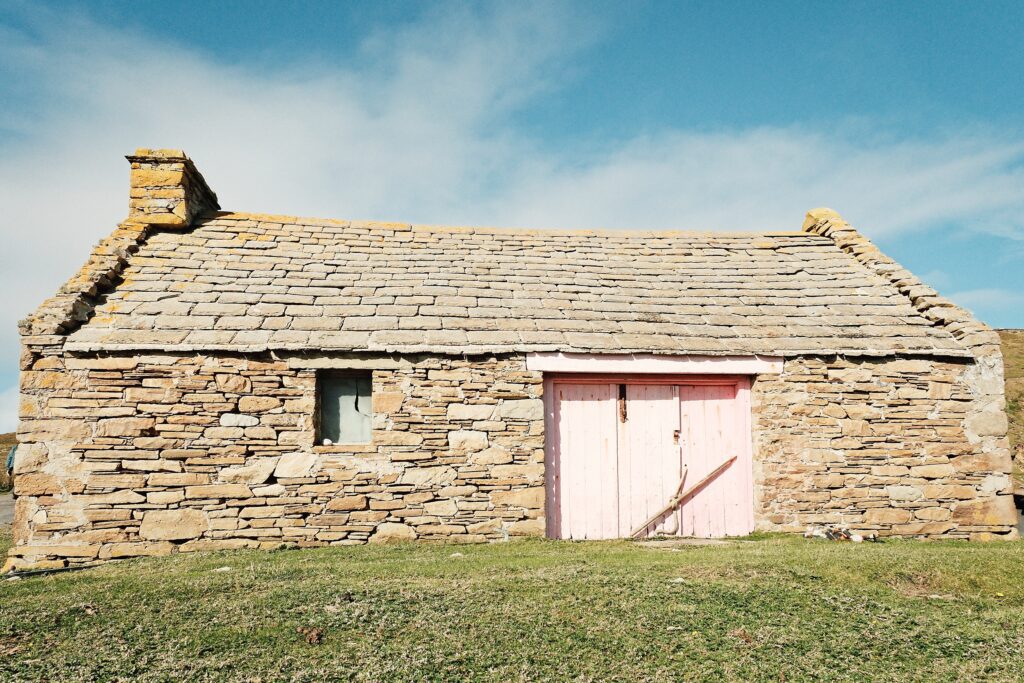 A minimalist architectural shot of a small stone croft house featuring a subtle pink door under a vast sky