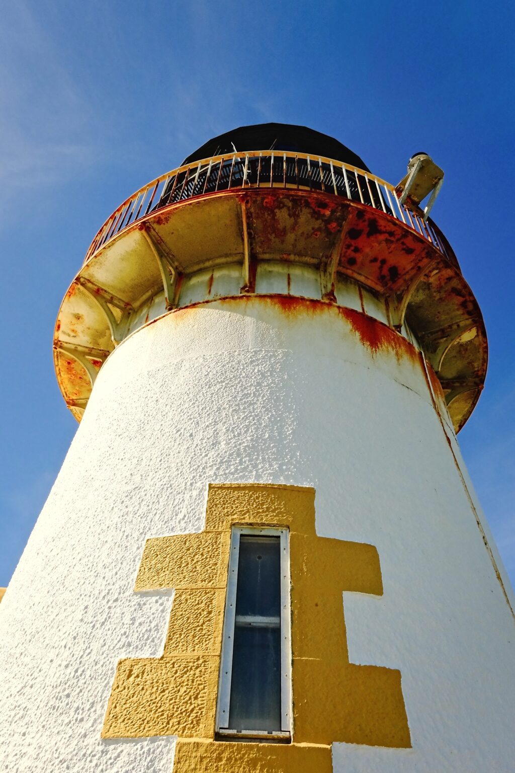 Architectural editorial shot of a weathered white and yellow lighthouse tower against a clear blue sky