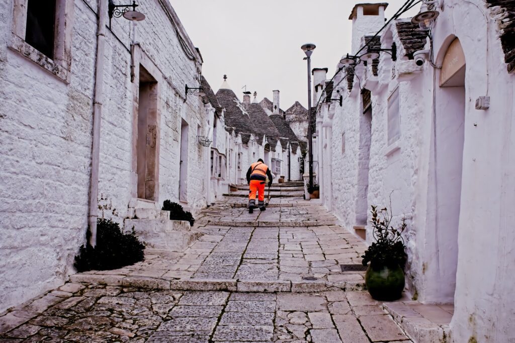 Honest documentary photography of a street cleaner in bright orange workwear, creating a sharp color contrast against the white-washed Trulli walls of Alberobello