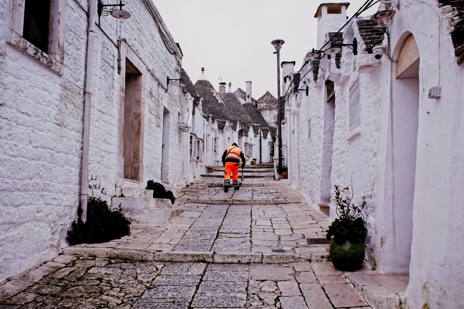 Honest documentary photography of a street cleaner in bright orange workwear, creating a sharp color contrast against the white-washed Trulli walls of Alberobello