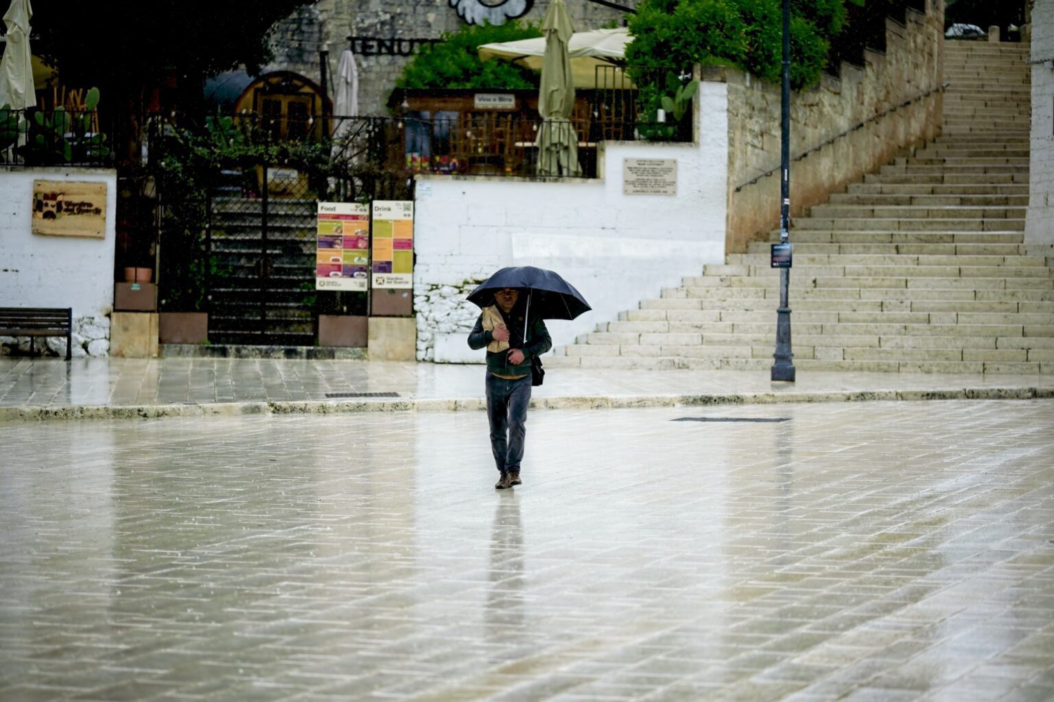 Candid street photography of a lone figure with a black umbrella crossing a rain-slicked limestone piazza in Puglia