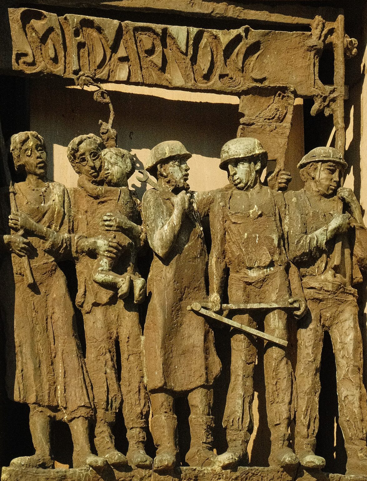 A bronze bas-relief sculpture titled "Solidarność" (Solidarity), depicting a group of shipyard workers, a woman, and a man holding a child, symbolizing the collective unity of the Polish labor movement.