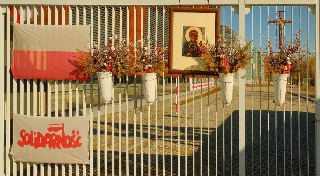 Close-up of the shipyard gate fence adorned with the Polish flag, a Solidarity banner, and floral tributes.