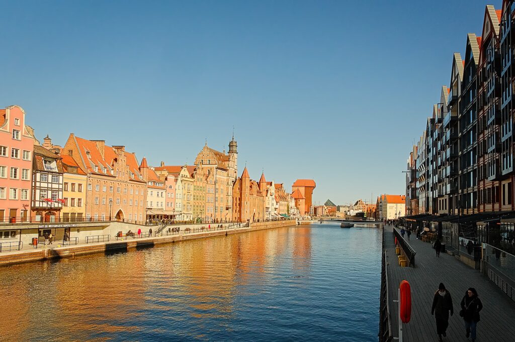 Cinematic wide-angle view of the Motława River canal in Gdańsk, featuring reflected Hanseatic architecture and the historic skyline under a clear sky.