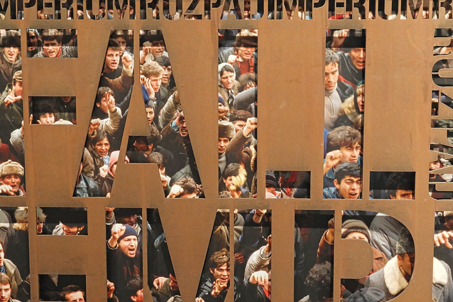 A close-up of a rusted metal commemorative panel with cut-out lettering revealing a dense crowd of protesters from the historic Polish shipyard strikes.