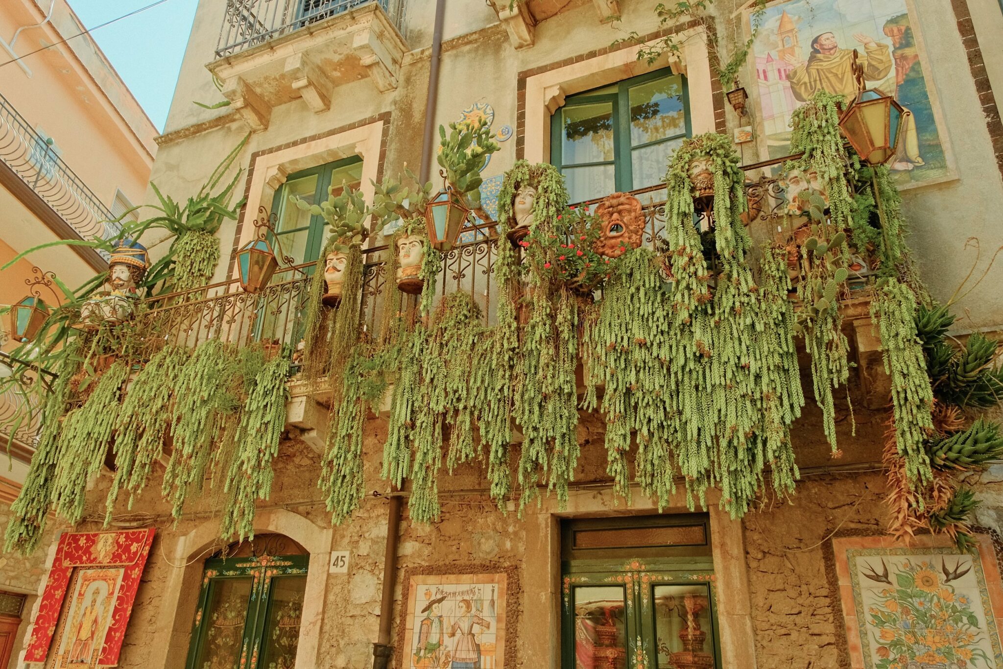Cinematic wide-angle architectural editorial of a historic Taormina building with dramatic greenery draping from ornate balconies