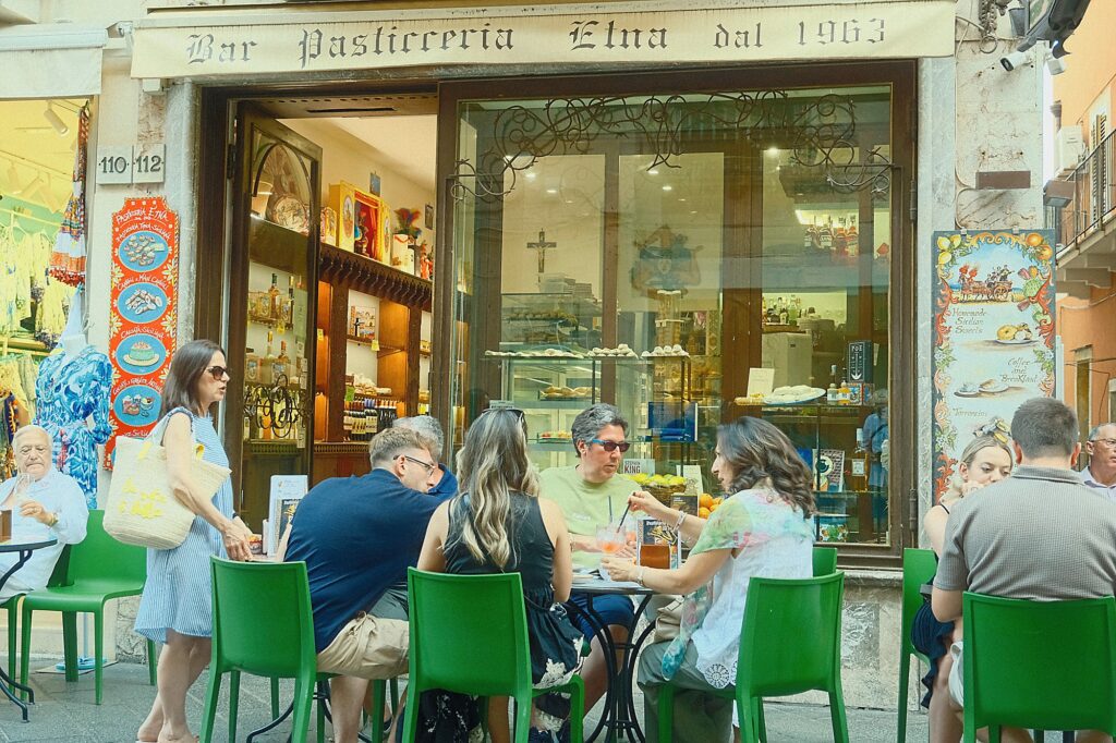 Candid documentary photography of people sitting at a green-tabled outdoor cafe in Taormina, Italy.