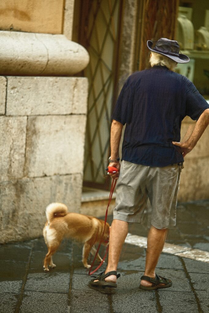 Unobtrusive street photography of a man walking his dog past a historic stone wall in a Sicilian side street