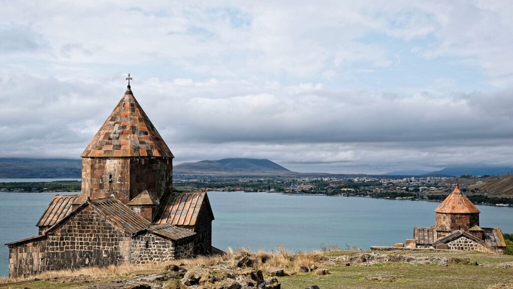 Wide-angle travel photography of an ancient stone monastery overlooking a calm lake and distant mountains in Georgia