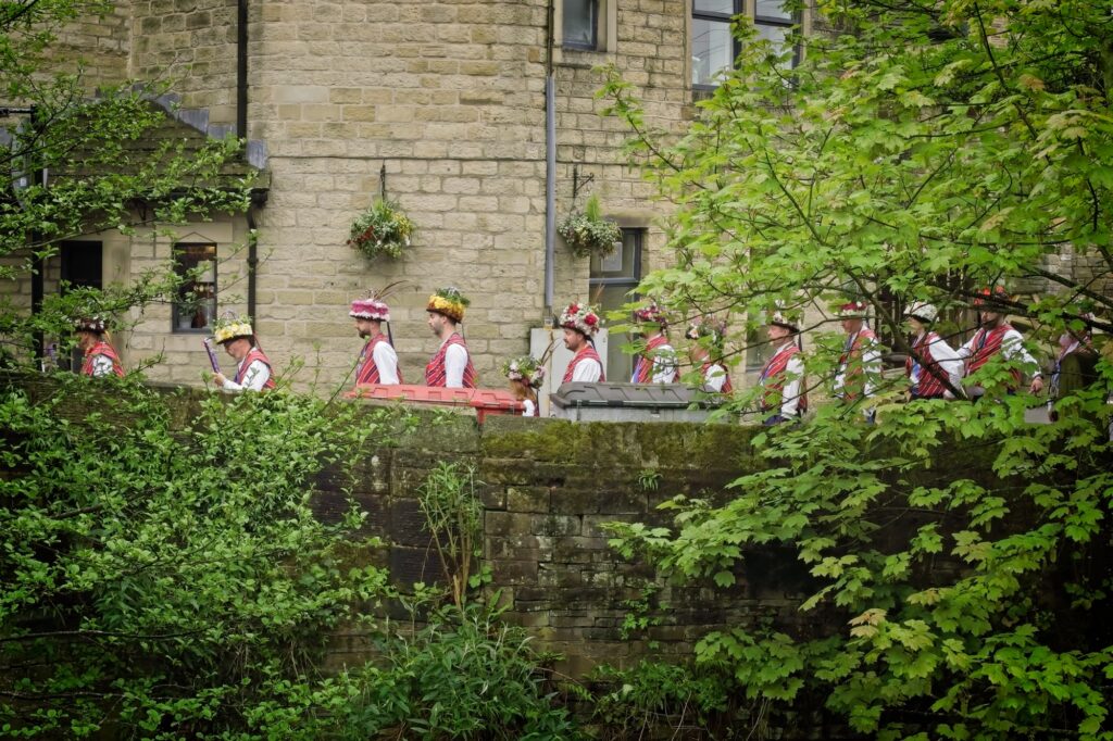 Cinematic observational photography of a traditional Morris dancing group waiting behind a stone wall and foliage at the Holmfirth Festival of Folk