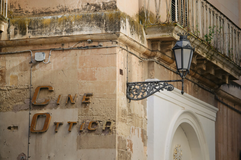 A weathered corner of an Italian building featuring a vintage "Cine Ottica" sign and an ornate wrought-iron street lamp against warm limestone.