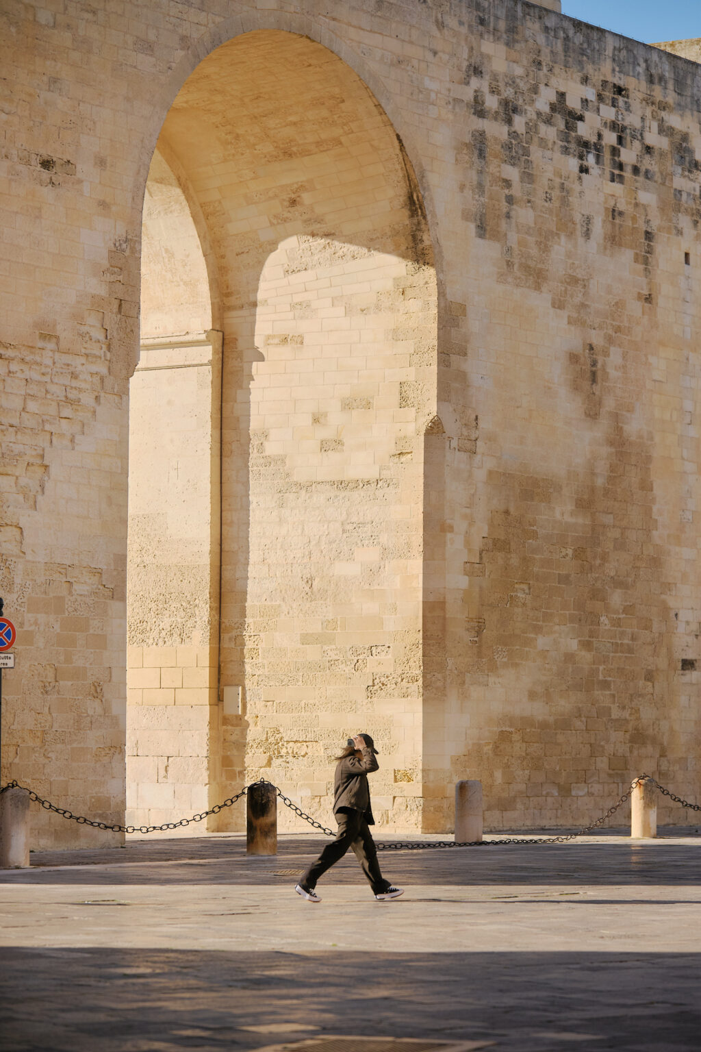 A person walking through the massive sun-drenched stone archway of Porta Napoli in Lecce, casting a long shadow across the historic paved square.
