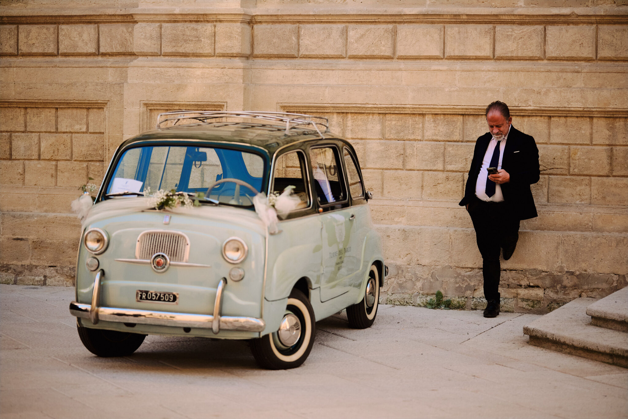 A vintage Fiat 600 Multipla decorated with flowers, parked in front of a textured stone wall with the driver in a suit leaning nearby.