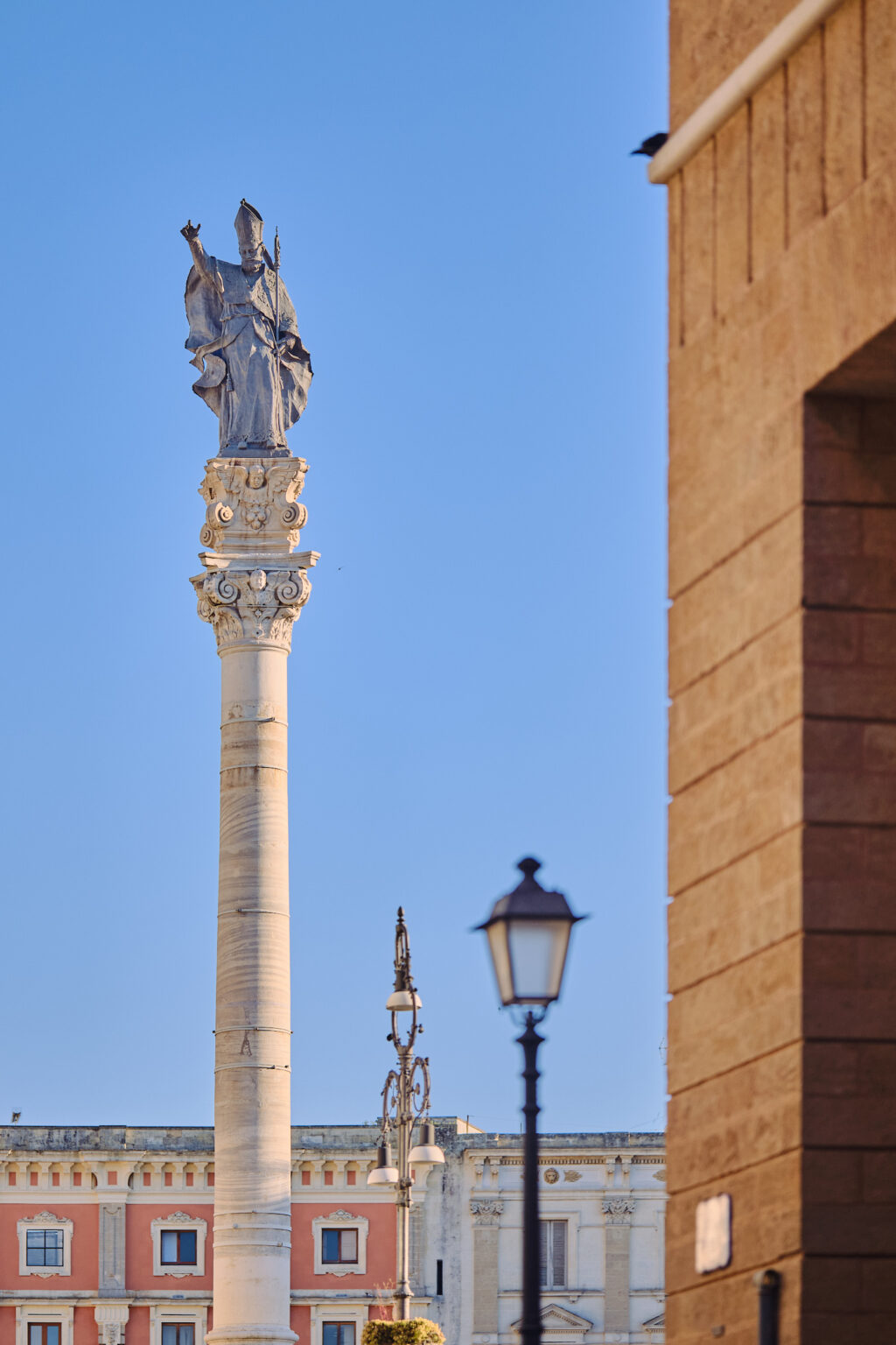 A tall stone column topped with a religious statue in Lecce, set against a vibrant blue sky with a classic street lamp in the foreground.