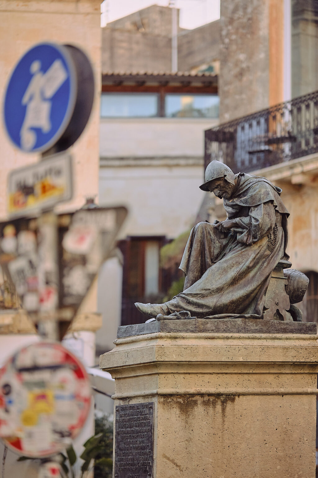 A bronze statue of a seated, contemplative figure in robes atop a stone pedestal, framed by out-of-focus street signs and historic buildings.