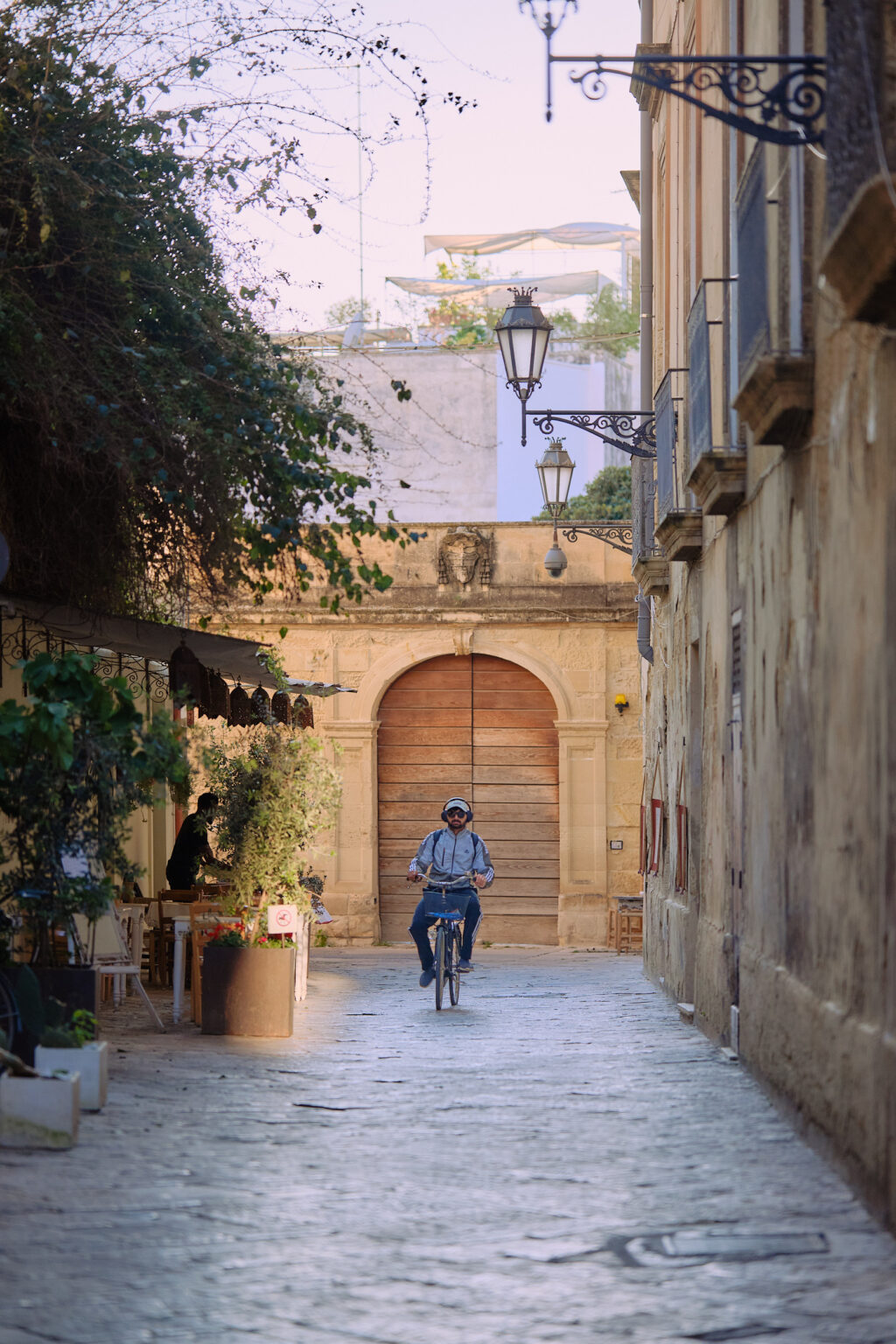 A cyclist riding down a narrow, atmospheric Lecce alleyway lined with greenery and cafe tables, leading toward a large wooden door.