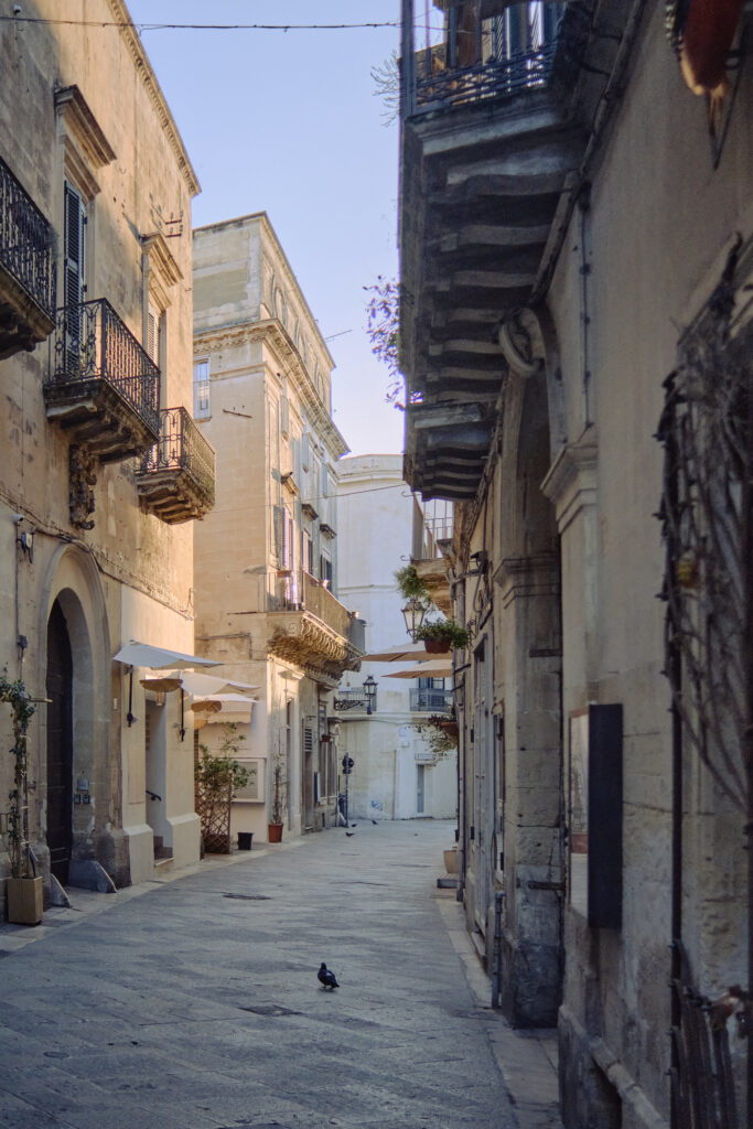 A quiet, sunlit Lecce street at dawn, with a single pigeon on the cobblestones and traditional balconies overlooking the path.