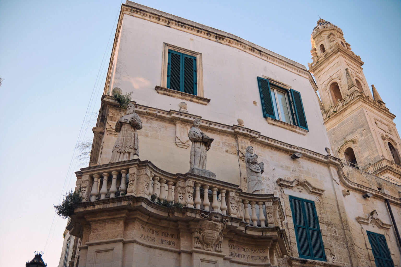 An elevated view of a historic building balcony adorned with stone statues, with a Baroque bell tower rising in the background.