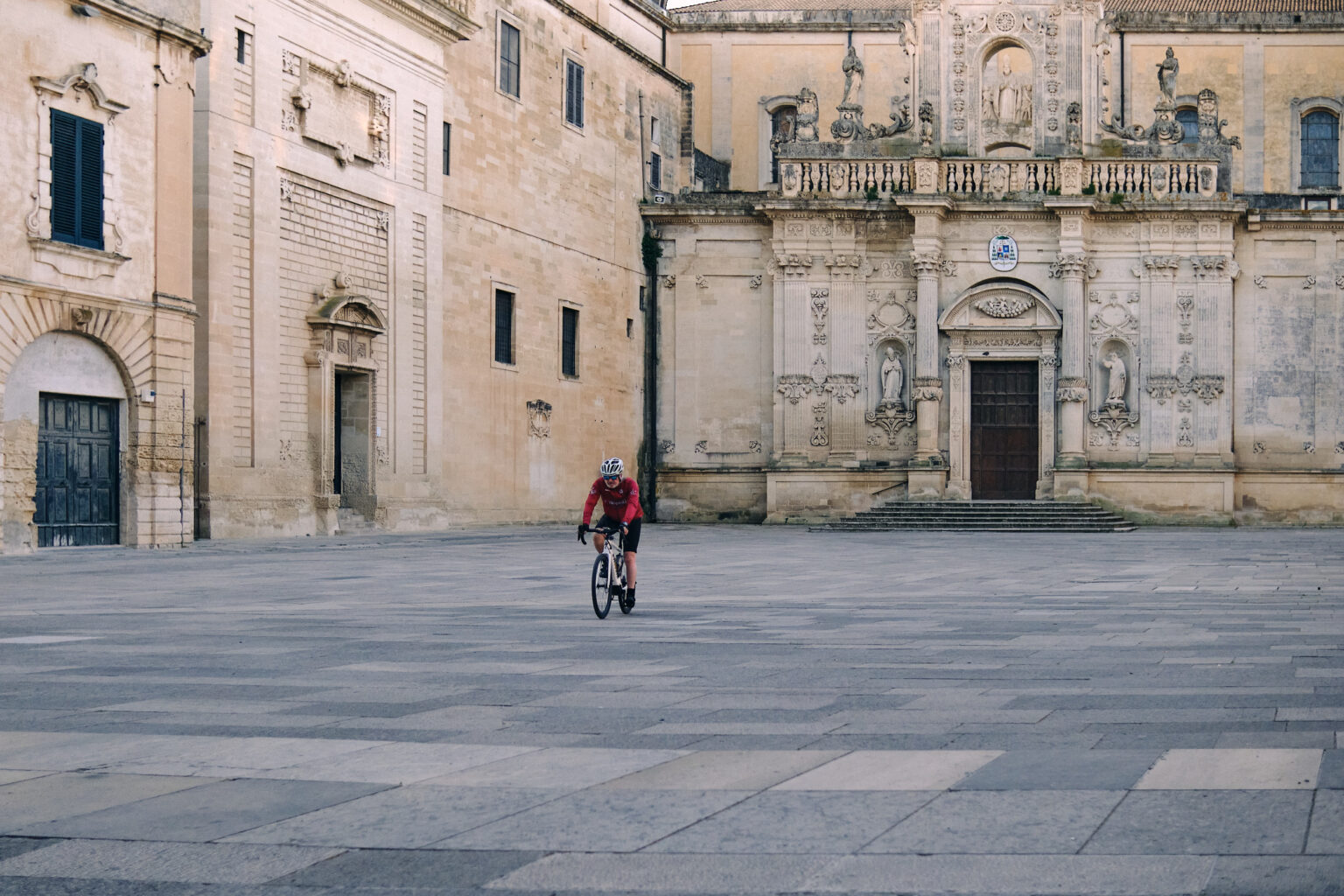 A wide-angle view of a vast, paved Italian piazza with a lone cyclist in red passing in front of an intricately carved cathedral facade.