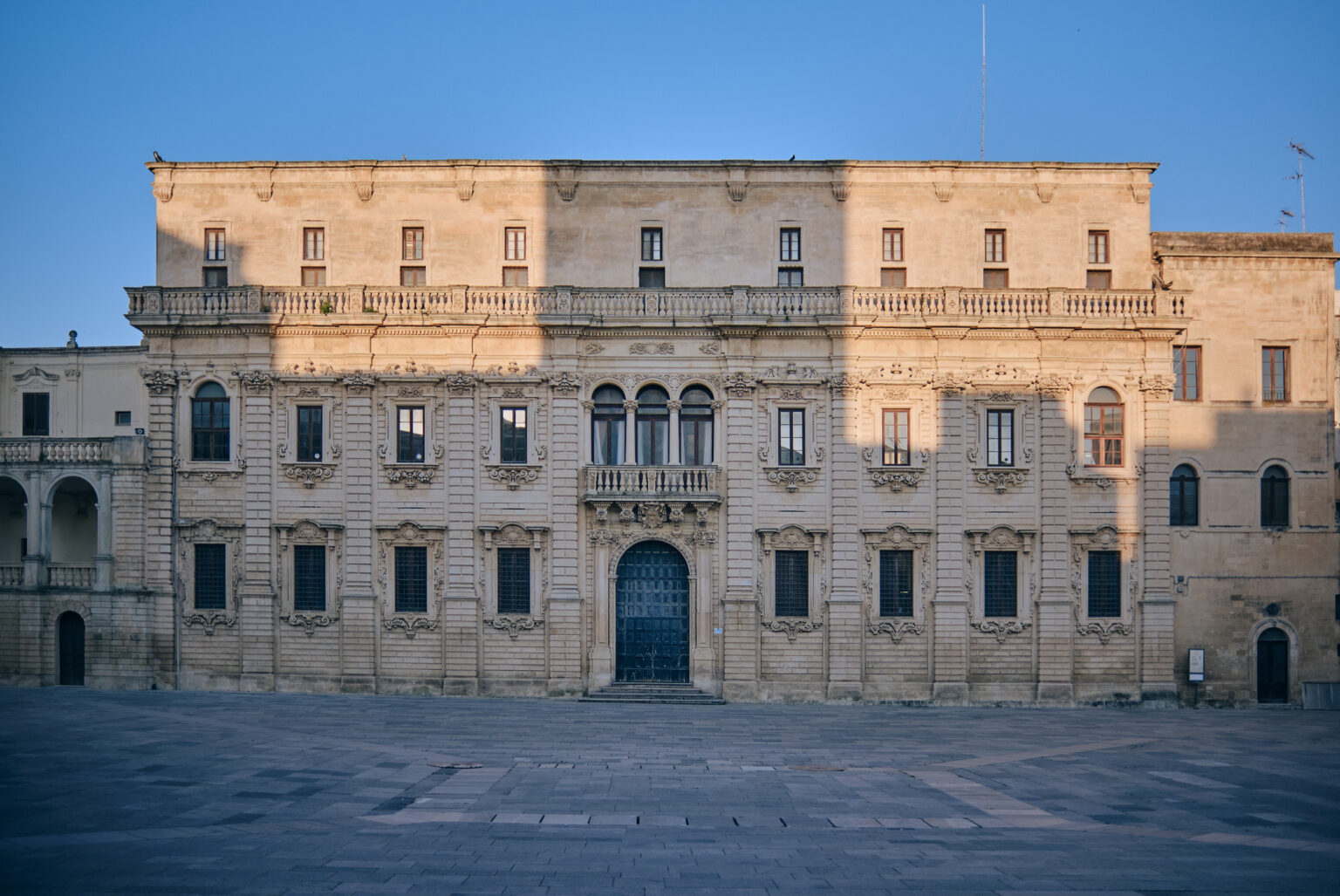 Wide architectural view of a grand Baroque palazzo in Lecce at dusk, featuring symmetrical windows and a central dark arched doorway under a deep blue sky.
