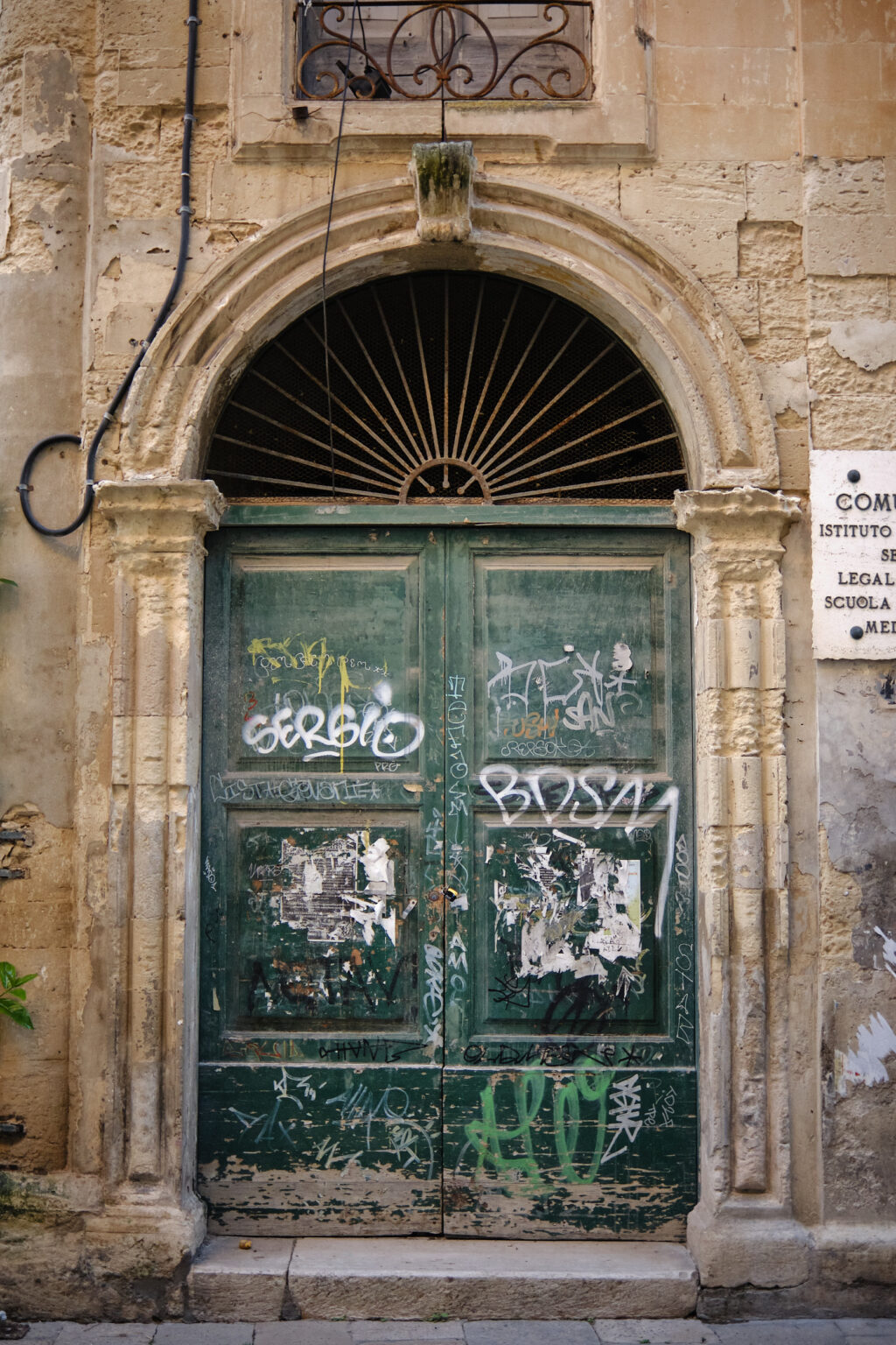 A distressed green wooden door with street art and graffiti, framed by a classic stone archway and ornate metal fanlight in an Italian alley.