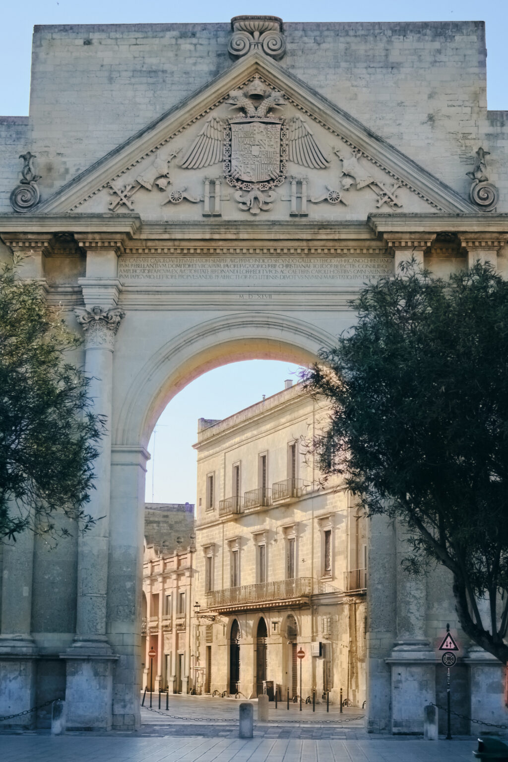 The majestic Porta Napoli triumphal arch in Lecce, shot through the archway toward sunlit historic buildings and street trees.