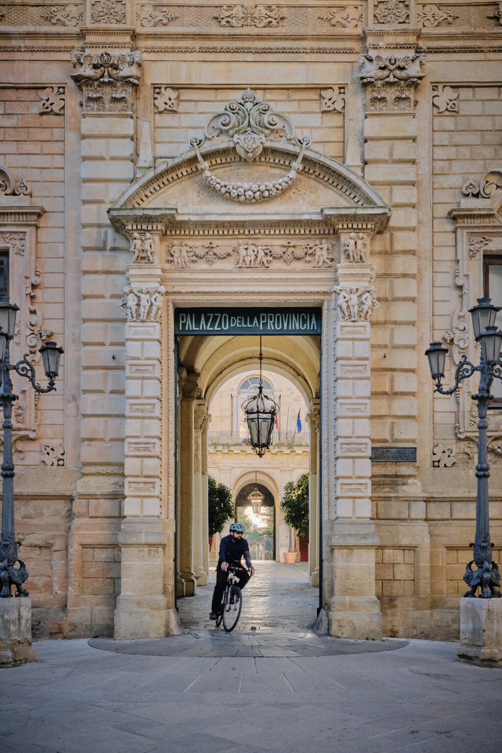 A lone cyclist passing through the grand arched entrance of the Palazzo della Provincia, framed by ornate Baroque stonework and hanging iron lanterns.