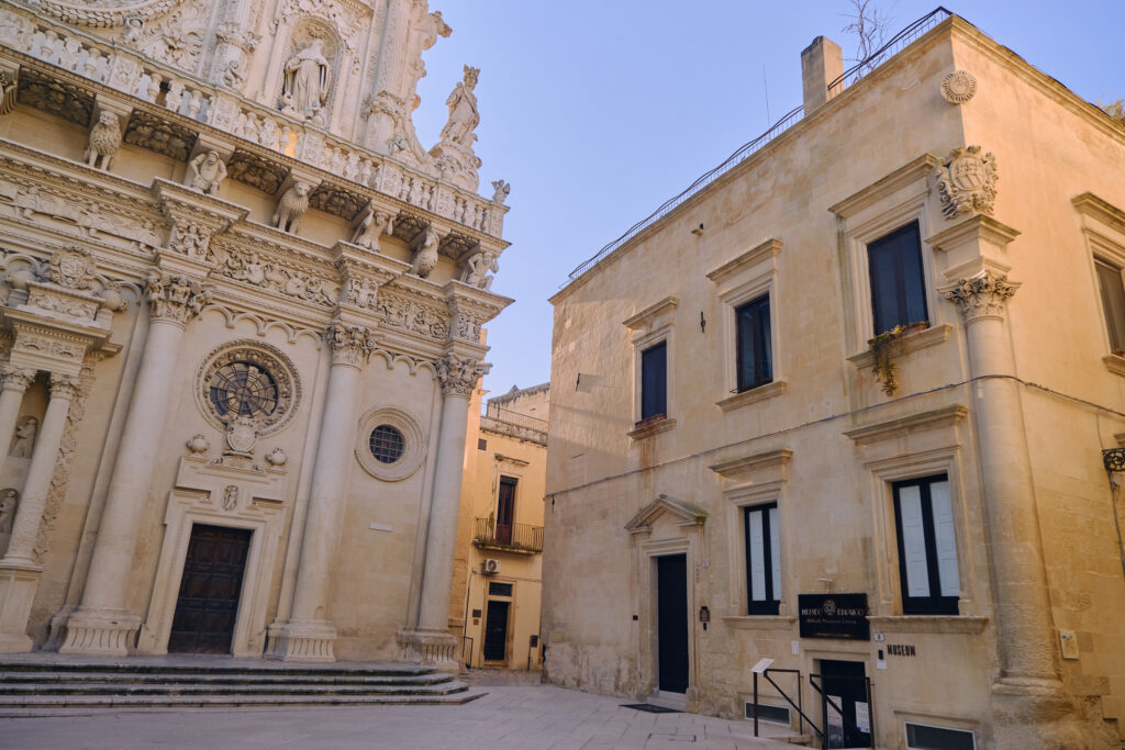 Perspective view of the intricate facade of the Santa Croce Basilica alongside the Jewish Museum Lecce under a clear sky.