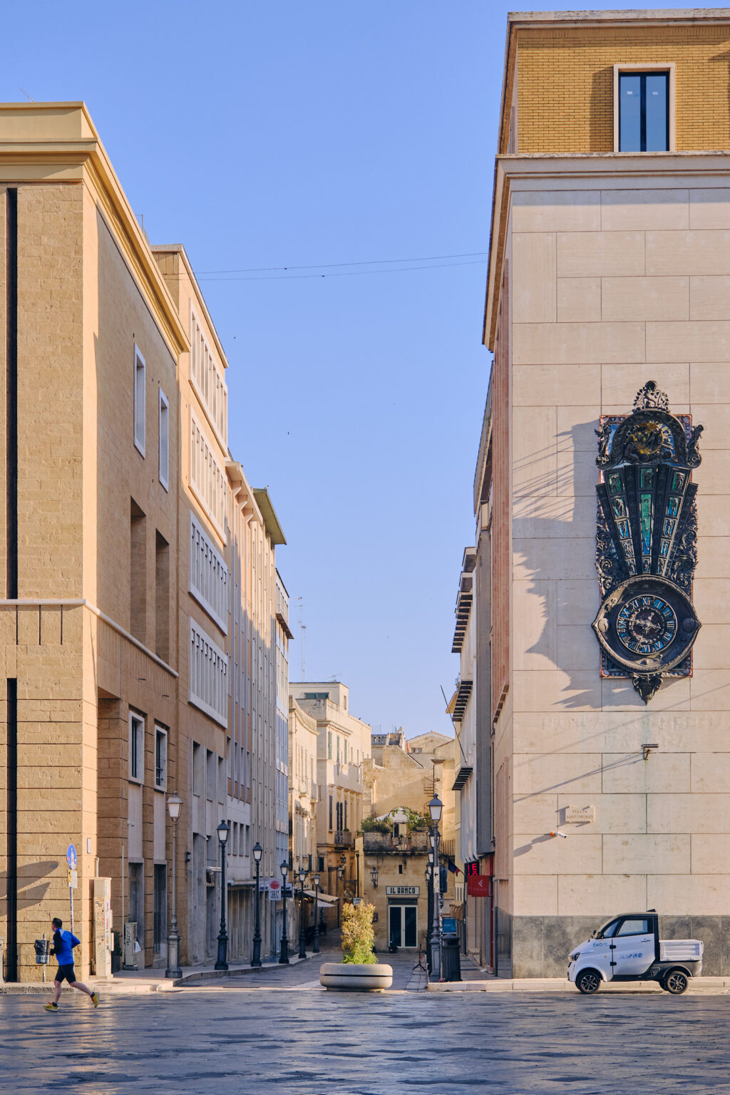 A modern street view in Lecce featuring a tall building with an ornate vertical clock and a small white electric utility vehicle parked in the foreground.