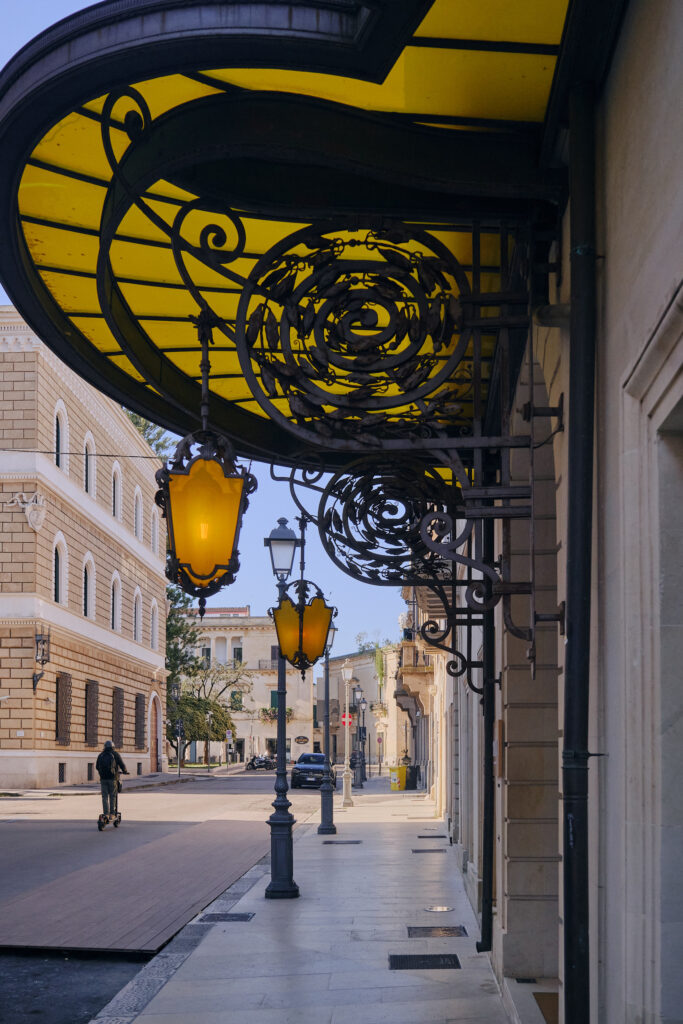 Dramatic low-angle shot of an Art Nouveau yellow glass canopy and decorative wrought-iron lanterns lining a quiet Lecce sidewalk.