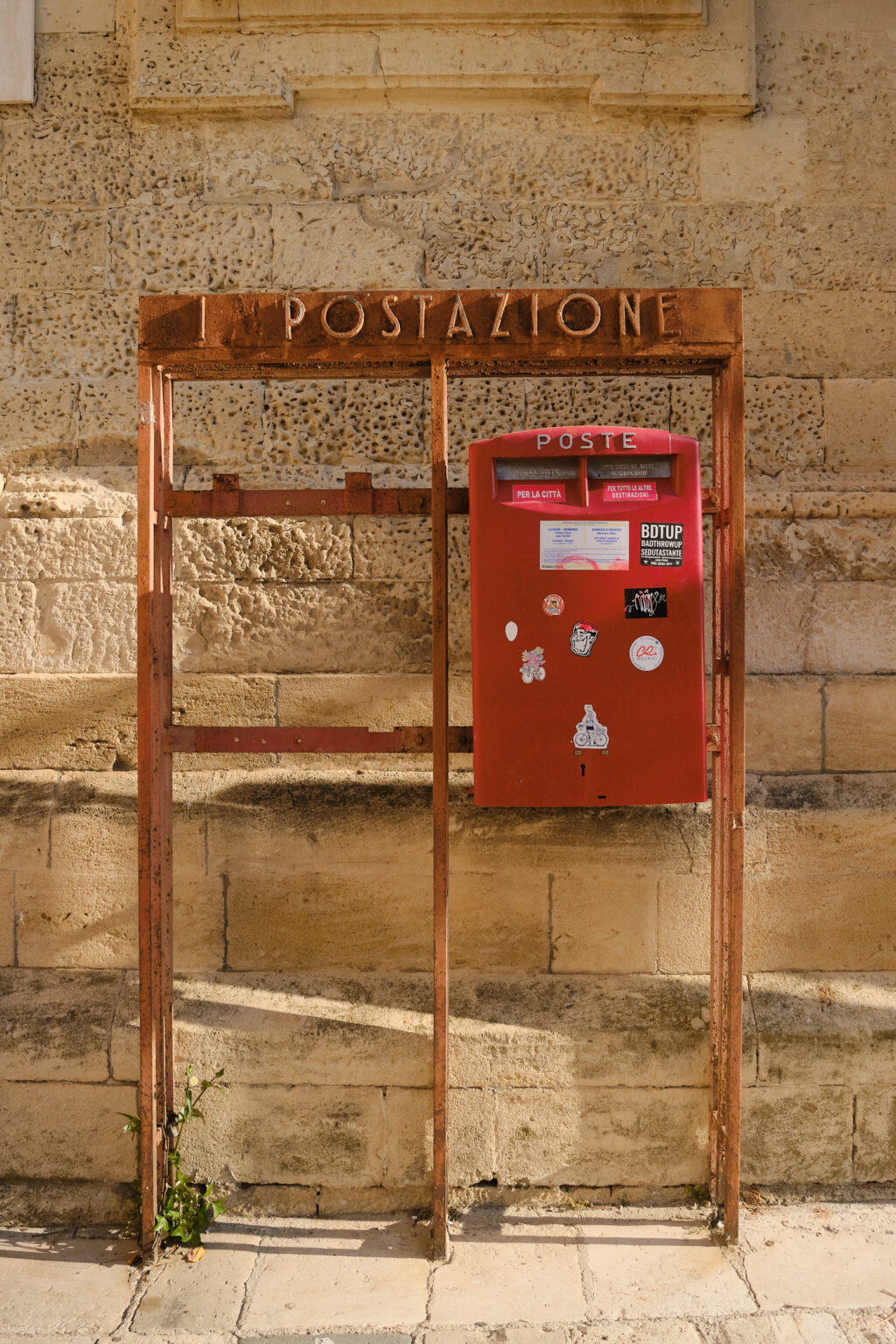 A weathered red Italian post box mounted on a rusted metal frame against a textured, sun-drenched limestone wall.