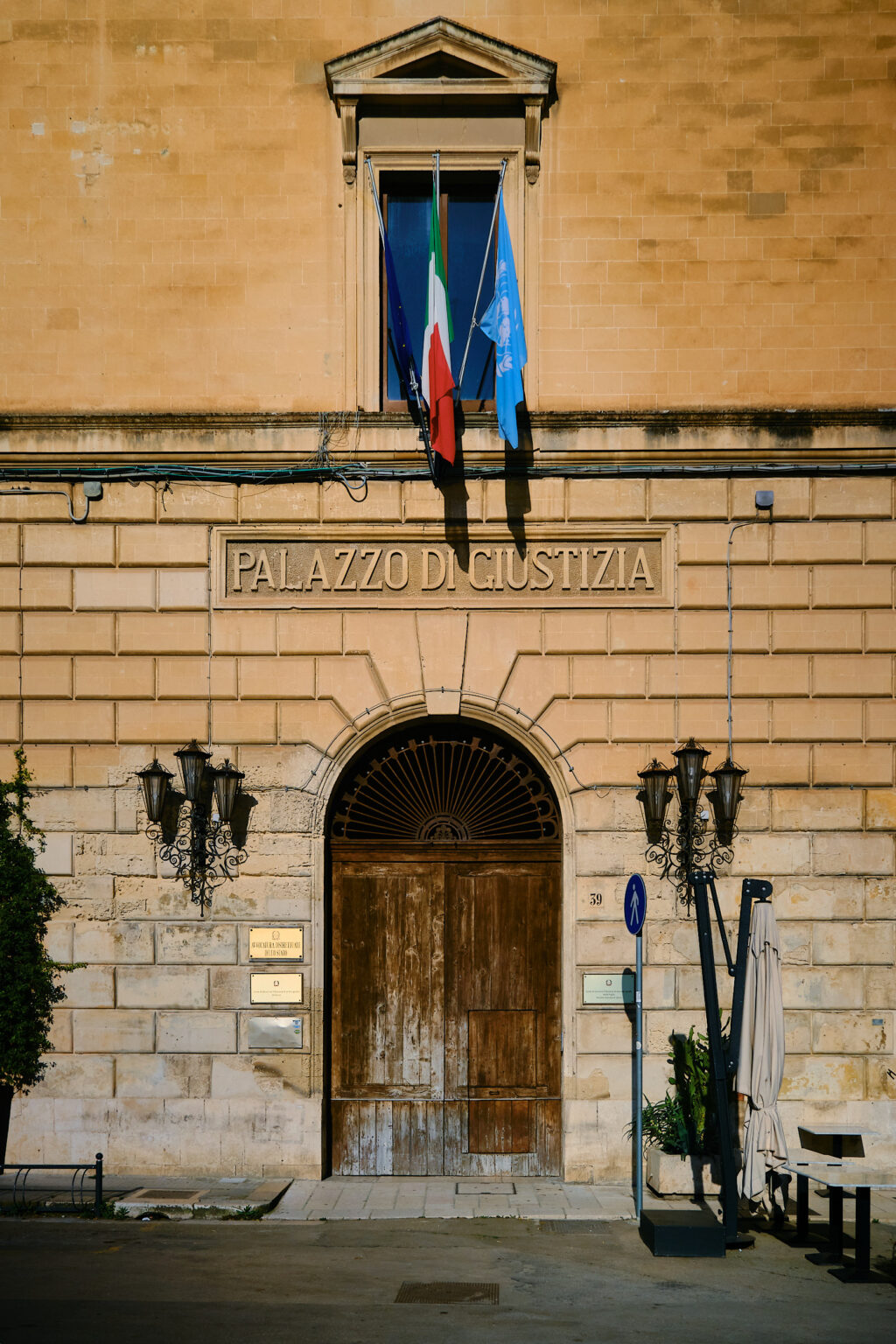The entrance to the Palazzo di Giustizia in Lecce, showing a massive wooden door, decorative iron lamps, and the Italian and European Union flags.