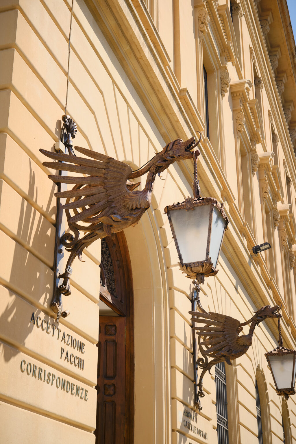 Ornate wrought-iron lanterns shaped like winged dragons or mythical creatures mounted on the side of a grand, golden-hued limestone building in Lecce, Italy, with historic signage for "Accettazione Pacchi" below.
