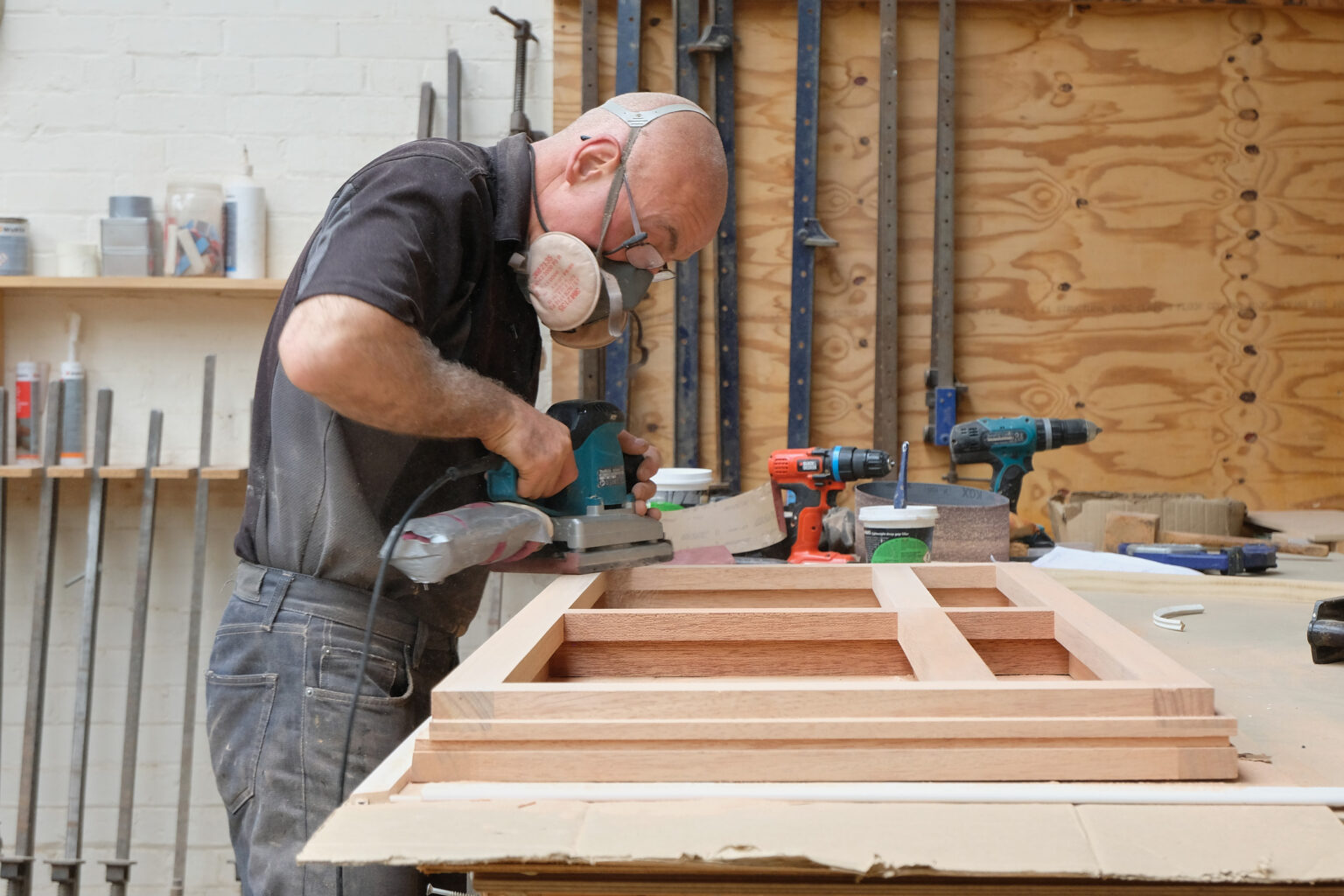 A carpenter wearing a dust mask using an electric sander on a custom wood frame in a joinery workshop.