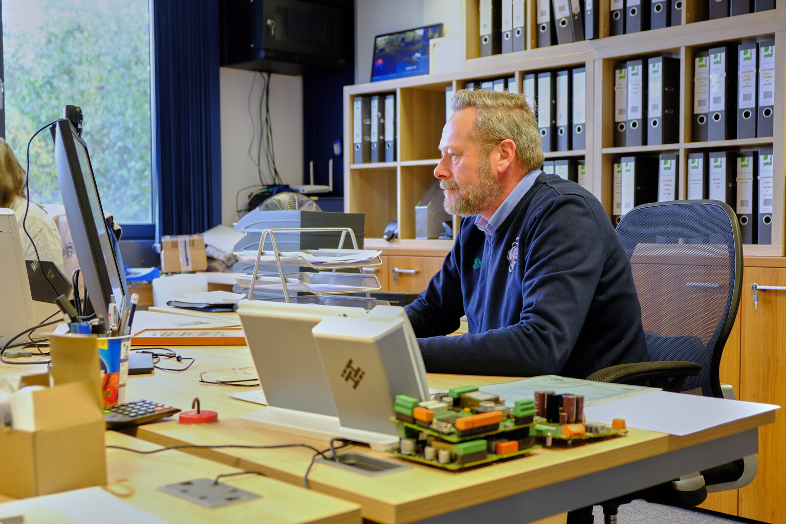 A professional male sitting at a large desk with hardware components in a clean, organized administrative workspace.