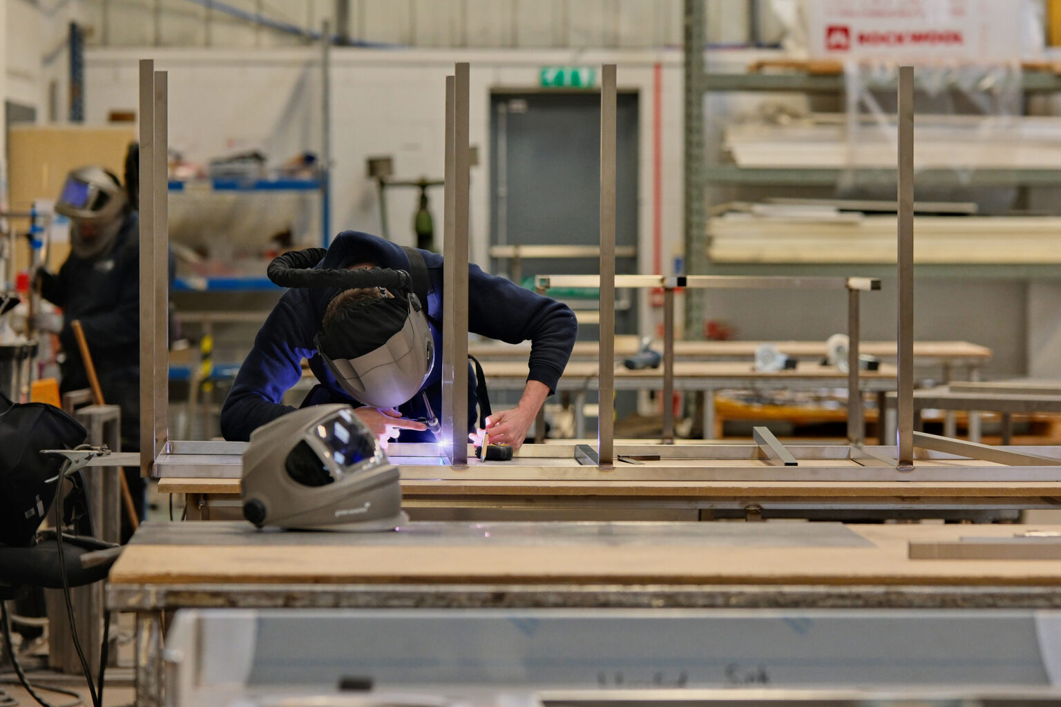 A fabricator welding metal components inside an industrial workshop, captured by a documentary photographer using selective focus.