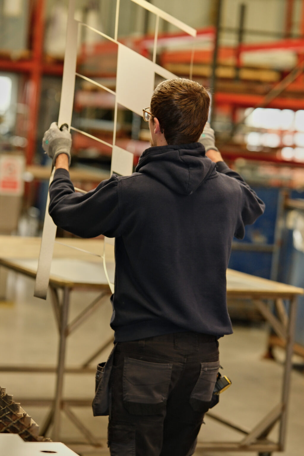An industrial workshop employee carrying a structural piece across a manufacturing floor.