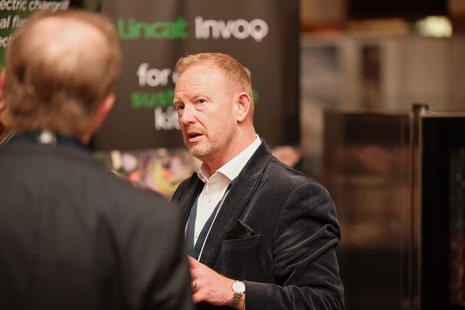 A male attendee in a black blazer talking to colleagues during a daytime corporate conference.