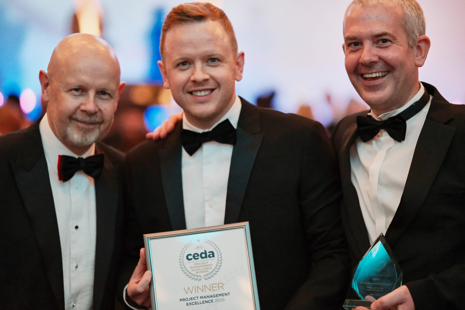 Three male colleagues in black tie attire holding up a framed award certificate at a business gala dinner.