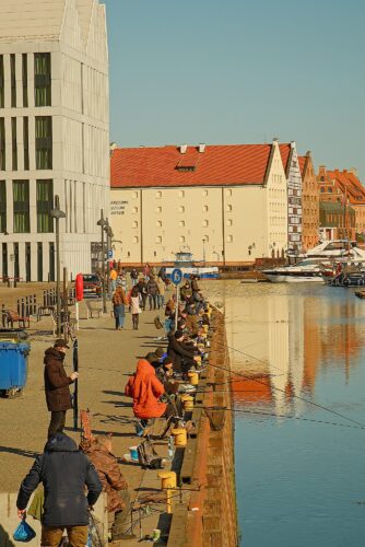 Documentary shot of local fishermen lined up along the concrete embankment of the Motława canal with modern architecture in the background.