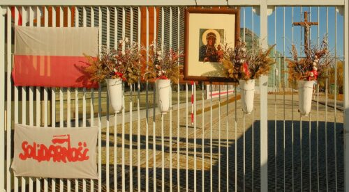 Close-up of the shipyard gate fence adorned with the Polish flag, a Solidarity banner, and floral tributes.