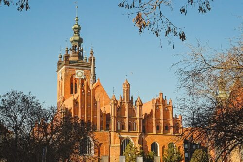 Low-angle view of the red-brick spires of St. Catherine’s Church, framed by tree branches against a blue sky.