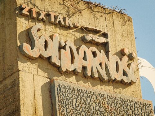 A low-angle shot of the wooden "21 Demands" plaques at the Gdańsk Shipyard, featuring the iconic "Solidarność" logo and text commemorating the August 1980 strikes.
