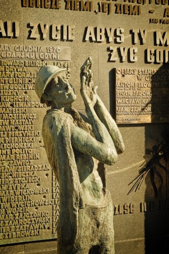 A bronze statue of a shipyard worker in a hard hat looking upward with hands raised, set against a memorial wall engraved with the names of the victims of the December 1970 protests.