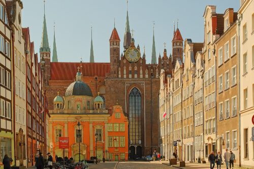An urban landscape looking down a historic cobblestone street toward the massive brick Gothic facade and astronomical clock of St. Mary's Church in the Gdańsk Old Town.